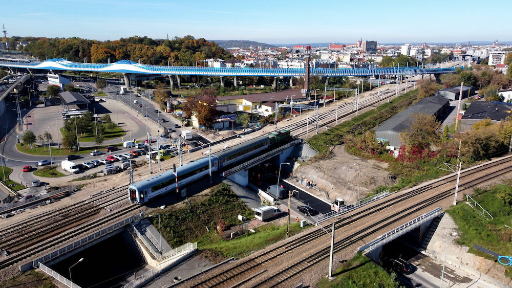 Zdjęcie przedstawia pociąg przejeżdżający na wiadukcie na ul. Powstańców Wielkopolskich w Krakowie. Pod wiaduktem kolejowym jest kładziony asfalt.