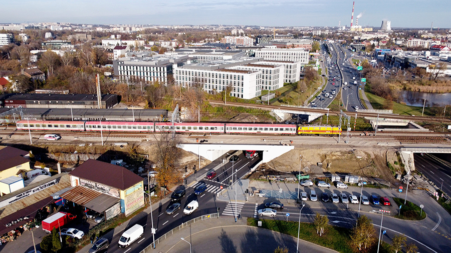 Zdjęcie przedstawia wiadukt na ulicy Dekerta w Krakowie. Zostało zrobione z lotu ptaka. Na zdjęciu widać jadący pociąg na wiadukcie oraz przejeżdżające samochody pod obiektem.
